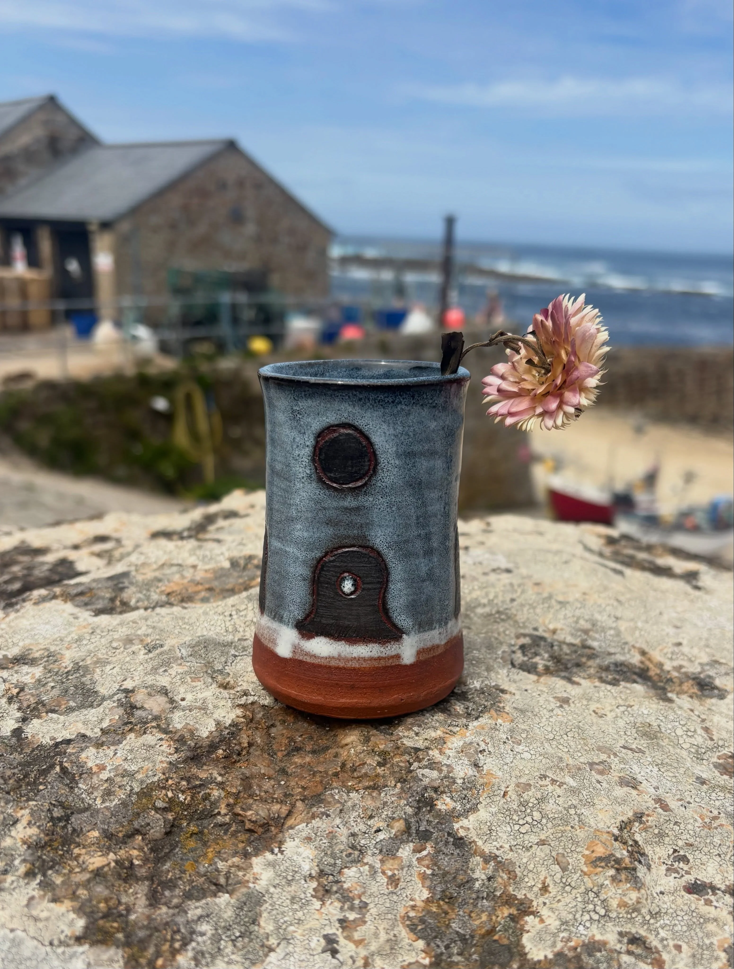 A blue-grey bud vase decorated with Mên an Tol standing stones, holding a pink flower beside the Cornish sea.
