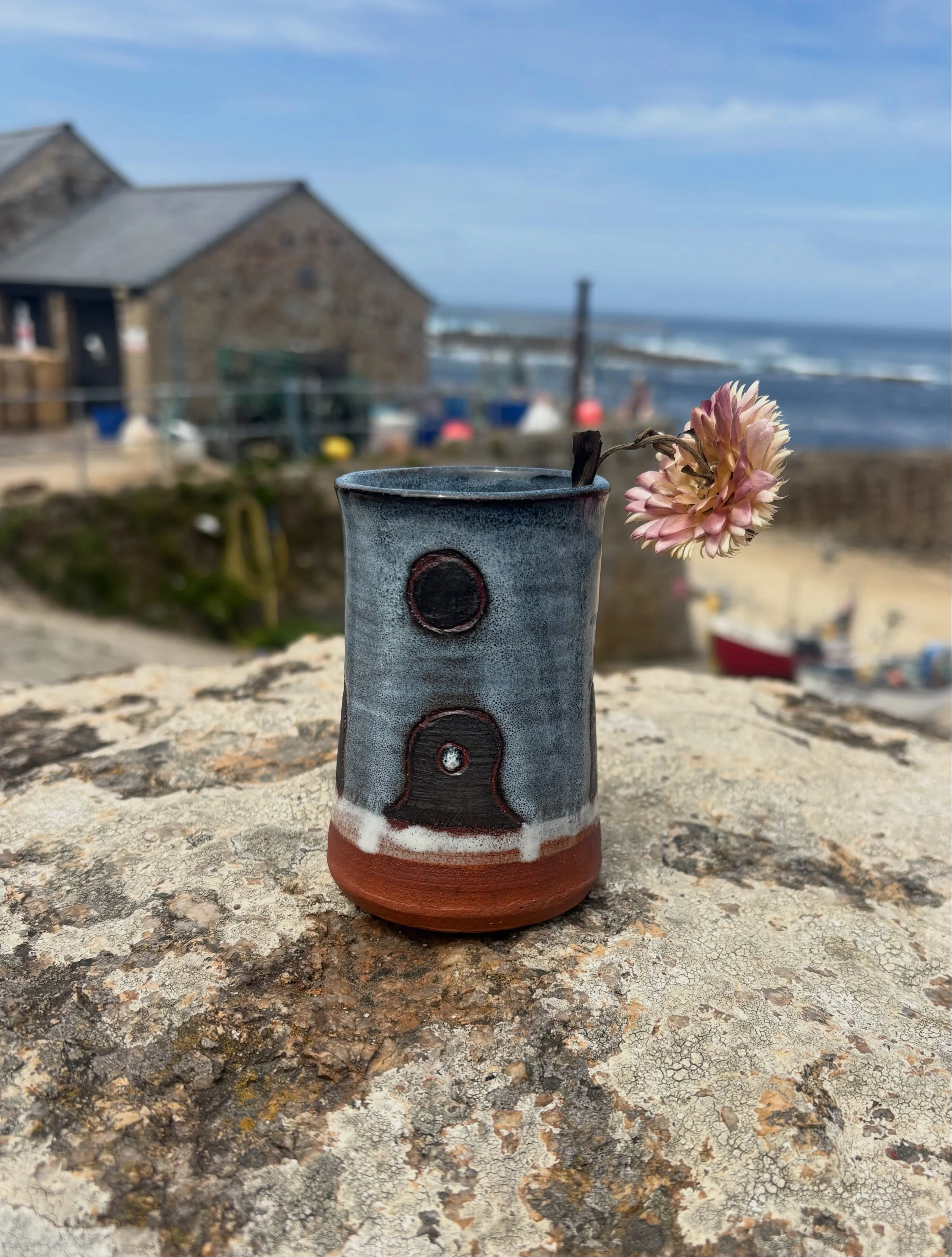 A blue-grey bud vase decorated with Mên an Tol standing stones, holding a pink flower beside the Cornish sea.