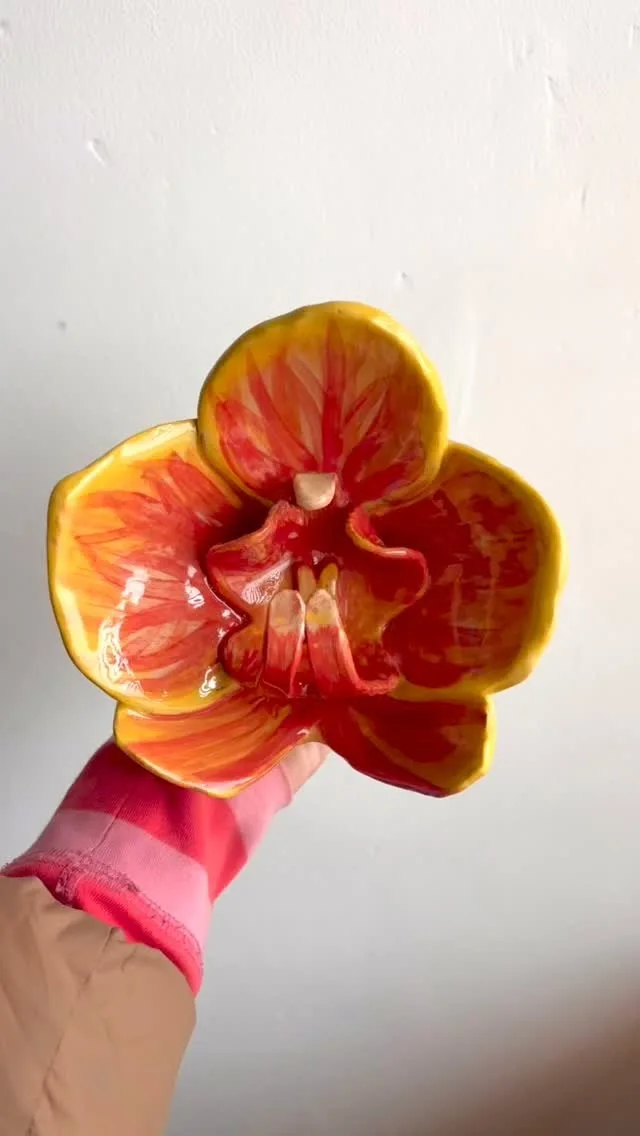 A glossy red and orange ceramic orchid with yellow petal edges, held in a hand against a pale wall.