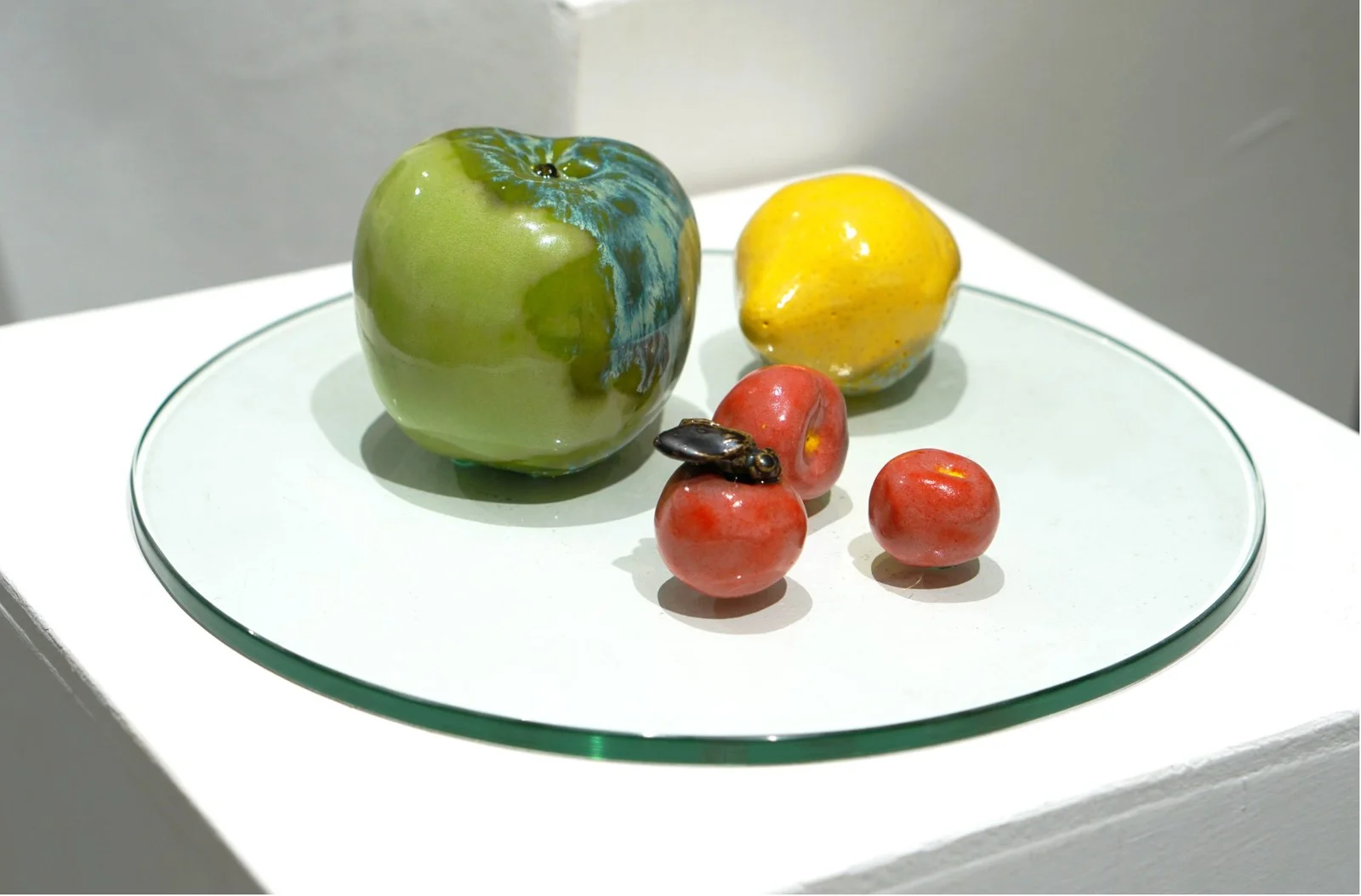 Ceramic green apple, yellow lemon and red cherries with a small insect, arranged on a round glass plate.