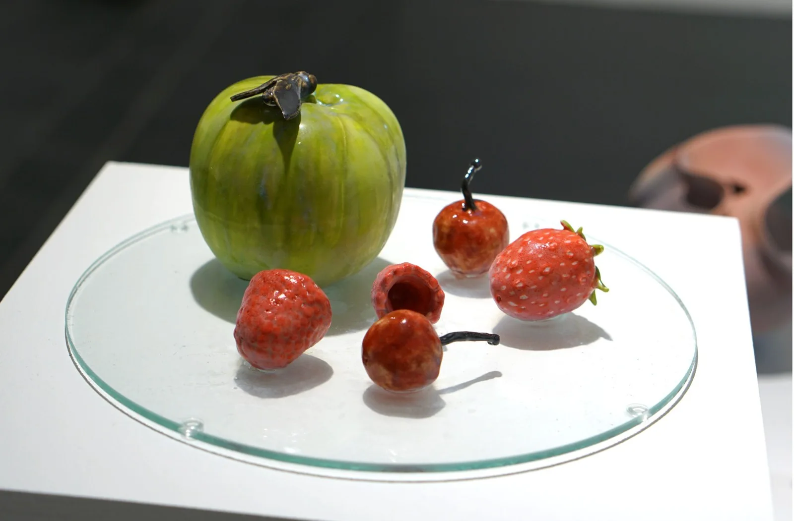 Green porcelain apple with an insect, red strawberries and cherries arranged on a clear glass plate.