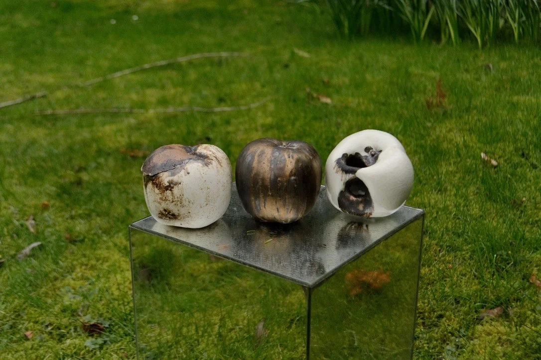 Three porcelain apple forms arranged on a mirrored plinth outdoors.