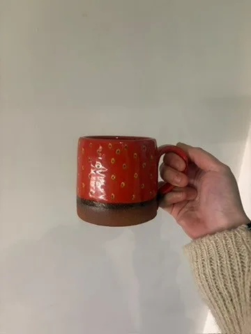 Red strawberry mug with small pale seed marks, a brown handle and an unglazed-looking base.