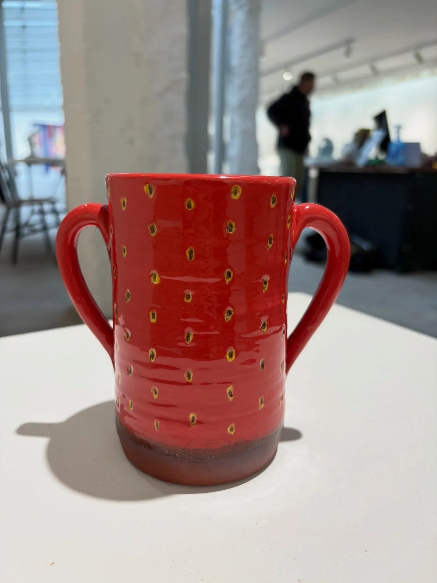 A tall red strawberry vase with two looping handles, small yellow seeds and a dark clay foot.