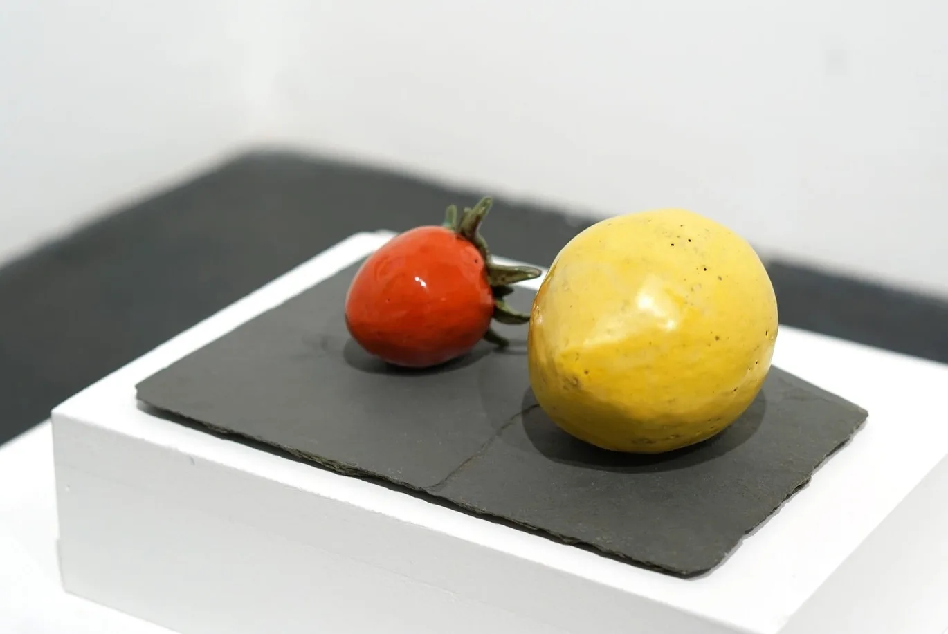 A red ceramic strawberry and yellow ceramic lemon resting together on a dark slate plinth.